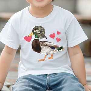May include: White t-shirt with a cartoon duck wearing a camouflage hat that says "Jaxon" on it. The duck is holding a red heart in its beak and there are three red hearts on the shirt.