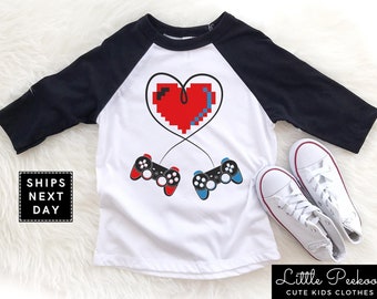 May include: White and black baseball tee shirt with a pixelated red heart and two video game controllers. The heart is connected to the controllers by cords. The shirt has the text "Ships Next Day" on the left side.