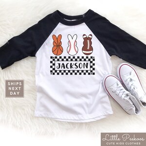 May include: White and black baseball tee shirt with a black and white checkered design and the name "JACKSON" printed on it. Three Easter bunny-shaped eggs are above the name, each decorated with a basketball, baseball, and football.