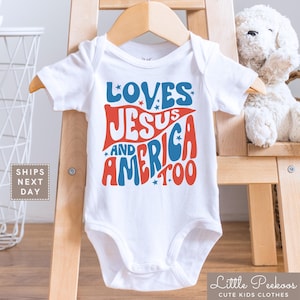 May include: White baby onesie with the text "LOVES JESUS AND AMERICA TOO" in red and blue. The onesie is displayed on a wooden hanger. The text is in a retro font with star accents. The onesie is part of the "Little Peekoos Cute Kids Clothes" collection.