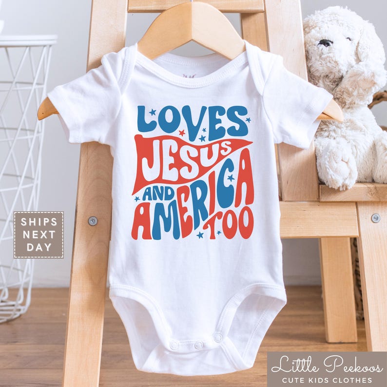 May include: White baby onesie with the text "LOVES JESUS AND AMERICA TOO" in red and blue. The onesie is displayed on a wooden hanger. The text is in a retro font with star accents. The onesie is part of the "Little Peekoos Cute Kids Clothes" collection.