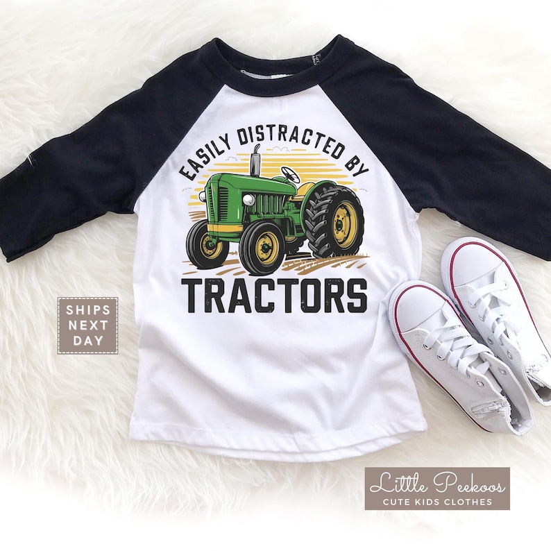 K&ouml;nnte beinhalten: Wei&szlig;es und schwarzes Raglan-&Auml;rmel-Shirt mit einer gr&uuml;nen und gelben Traktorgrafik. Das Shirt tr&auml;gt den Text "EASILY DISTRACTED BY TRACTORS". Wei&szlig;e Turnschuhe liegen neben dem Shirt.
