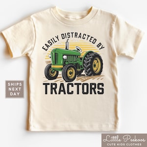 K&ouml;nnte beinhalten: Cremefarbenes T-Shirt f&uuml;r Kleinkinder mit einer gr&uuml;nen und gelben Traktorgrafik. Der Text &uuml;ber dem Traktor lautet "EASILY DISTRACTED BY" und der Text darunter lautet "TRACTORS". Das Shirt ist von Little Peekoos.