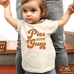 May include: A white toddler t-shirt with a brown graphic that says "Pies Before Guys" in a retro font.