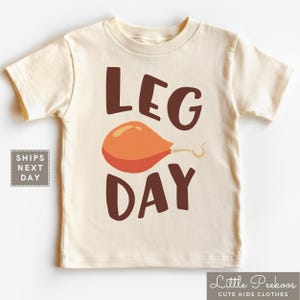 May include: A beige short-sleeved t-shirt with the text "LEG DAY" in brown letters. A cartoon orange turkey leg is in the middle of the text.