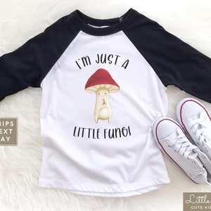 May include: White and black baseball tee shirt with a red and white mushroom graphic and the text "I'm just a little fungi".