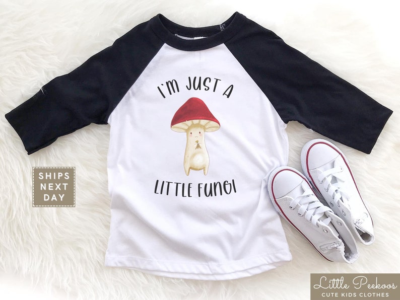 May include: White and black baseball tee shirt with a red and white mushroom graphic and the text "I'm just a little fungi".