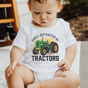 K&ouml;nnte beinhalten: Wei&szlig;er Baby-Body mit einem gr&uuml;nen und gelben Traktorgrafik. Der Text auf dem Body lautet "EASILY DISTRACTED BY TRACTORS". Das Traktormotiv zeigt einen gr&uuml;nen Traktor mit gelben R&auml;dern und einer gelben Sonne.