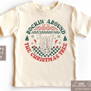 May include: A cream colored t-shirt with a red circle graphic that says "Rockin' Around The Christmas Tree". The graphic features a skeleton hand making a rock and roll sign, a checkered pattern, and a Christmas tree.