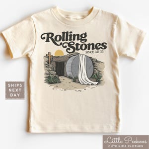 May include: A beige short-sleeved t-shirt with a graphic of a stone tomb with a white sheet draped over it. The text "Rolling Stones Since AD 33" is above the tomb.
