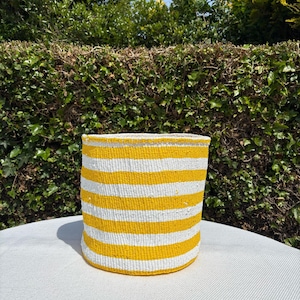May include: A cylindrical woven basket featuring a repeating pattern of horizontal yellow and white stripes. The basket has a flat base and an open top, constructed from a textured material with a consistent, tight weave throughout the structure.