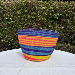 May include: A hand-woven basket featuring horizontal stripes in vibrant neon pink, bright yellow, turquoise, and deep purple. The basket has a wide, open top and a tapered, rounded base, constructed from tightly coiled, colourful natural fibres.