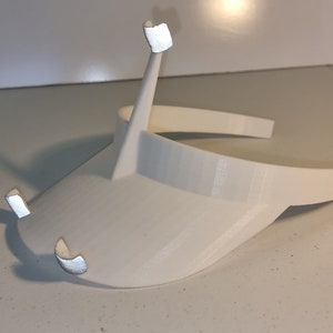 Puede incluir: Visera de plástico blanco con dos pequeños brazos de plástico blanco curvos que se extienden desde los lados. La visera está diseñada para ser usada en la cabeza.