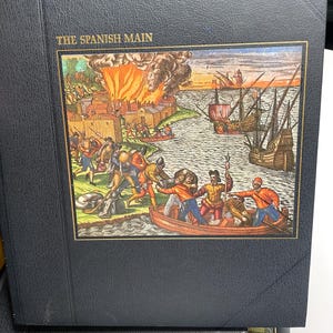 Puede incluir: Un libro negro titulado "THE SPANISH MAIN" con una colorida ilustraci&oacute;n que representa una escena de batalla hist&oacute;rica. La imagen muestra barcos, una ciudad en llamas y figuras en botes y en tierra, sugiriendo un tema hist&oacute;rico o n&aacute;utico.