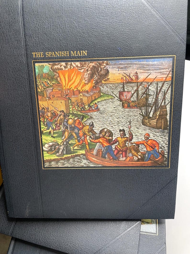 Puede incluir: Un libro negro titulado "THE SPANISH MAIN" con una colorida ilustraci&oacute;n que representa una escena de batalla hist&oacute;rica. La imagen muestra barcos, una ciudad en llamas y figuras en botes y en tierra, sugiriendo un tema hist&oacute;rico o n&aacute;utico.