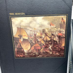 Puede incluir: Un libro azul oscuro titulado "THE ARMADA" con un borde dorado alrededor de una pintura hist&oacute;rica de barcos en el mar. La imagen representa una batalla naval con barcos, banderas y cielos tormentosos. El libro tiene una cubierta texturizada.