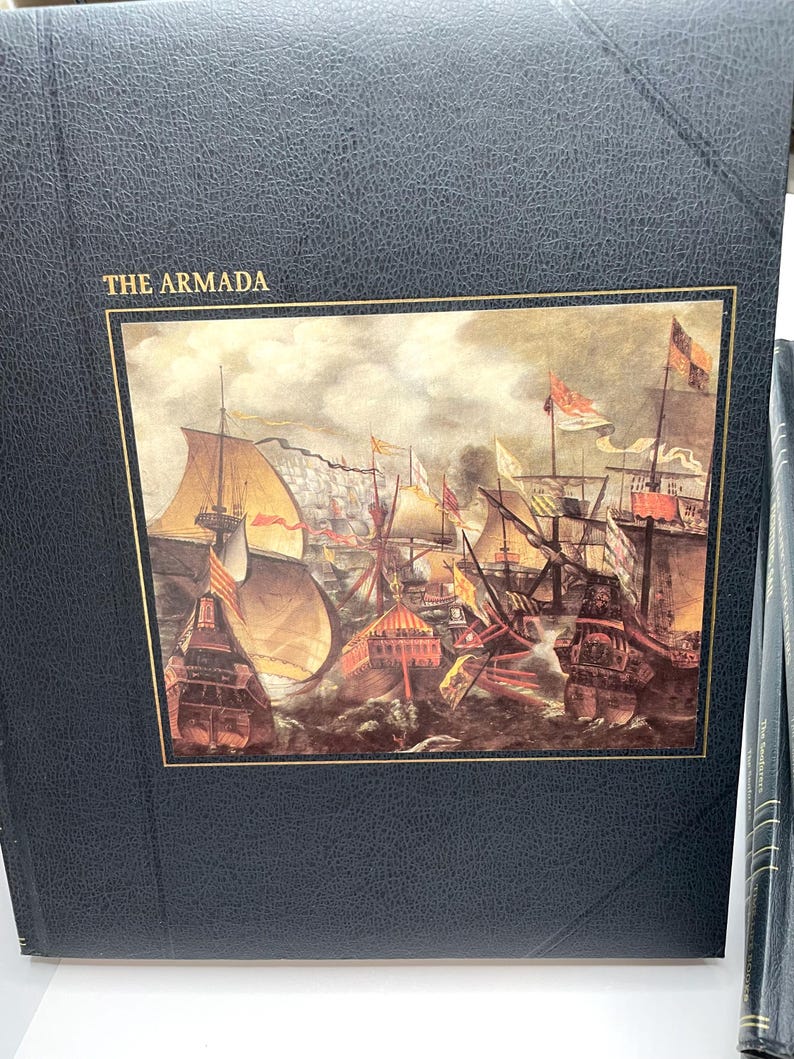 Puede incluir: Un libro azul oscuro titulado "THE ARMADA" con un borde dorado alrededor de una pintura hist&oacute;rica de barcos en el mar. La imagen representa una batalla naval con barcos, banderas y cielos tormentosos. El libro tiene una cubierta texturizada.