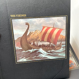 Puede incluir: &Aacute;lbum negro con ribete dorado y el t&iacute;tulo "THE VIKINGS". La portada presenta una ilustraci&oacute;n detallada de un barco vikingo navegando en un mar agitado con una vela a rayas rojas y blancas. Una cordillera se encuentra en el fondo.