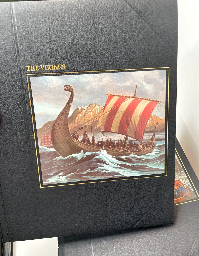 Puede incluir: &Aacute;lbum negro con ribete dorado y el t&iacute;tulo "THE VIKINGS". La portada presenta una ilustraci&oacute;n detallada de un barco vikingo navegando en un mar agitado con una vela a rayas rojas y blancas. Una cordillera se encuentra en el fondo.