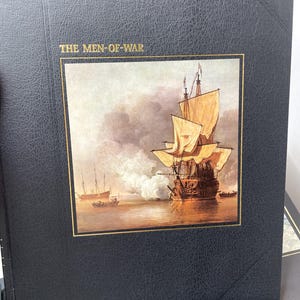 Puede incluir: Un libro negro titulado "THE MEN-OF-WAR" con un borde dorado alrededor de una pintura de un barco alto en el agua. El barco tiene velas de color canela y est&aacute; disparando un ca&ntilde;&oacute;n. Otros barcos est&aacute;n en la distancia.