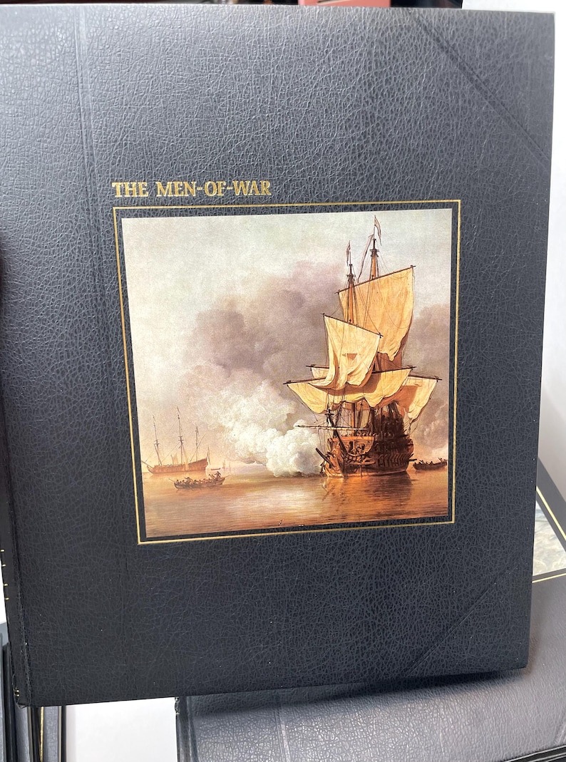Puede incluir: Un libro negro titulado "THE MEN-OF-WAR" con un borde dorado alrededor de una pintura de un barco alto en el agua. El barco tiene velas de color canela y est&aacute; disparando un ca&ntilde;&oacute;n. Otros barcos est&aacute;n en la distancia.