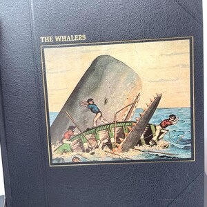 Puede incluir: Un libro azul oscuro titulado "THE WHALERS" con una ilustraci&oacute;n enmarcada de una ballena atacando un peque&ntilde;o bote. La ilustraci&oacute;n muestra a hombres en el bote, con la ballena rompiendo el agua. El libro tiene un borde dorado.