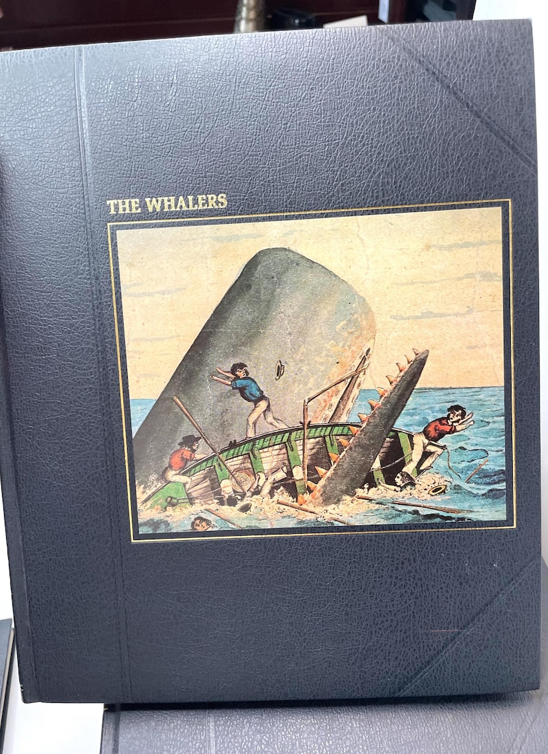 Puede incluir: Un libro azul oscuro titulado "THE WHALERS" con una ilustraci&oacute;n enmarcada de una ballena atacando un peque&ntilde;o bote. La ilustraci&oacute;n muestra a hombres en el bote, con la ballena rompiendo el agua. El libro tiene un borde dorado.