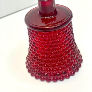 May include: A deep red glass bell featuring a textured hobnail pattern across its exterior surface. The bell has a smooth, flat top handle and a flared base, crafted from translucent ruby-colored glass that catches the light.
