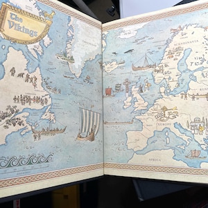 Puede incluir: Libro abierto con un mapa detallado del mundo vikingo. El mapa incluye ilustraciones de barcos vikingos, asentamientos y regiones geogr&aacute;ficas, como Am&eacute;rica del Norte, Europa y Escandinavia. El t&iacute;tulo "The Vikings" es visible en la parte superior.