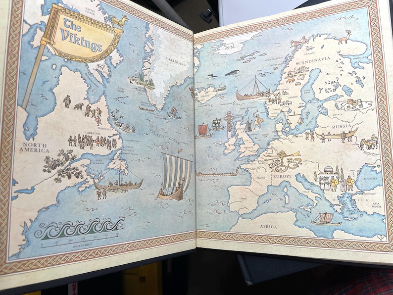 Puede incluir: Libro abierto con un mapa detallado del mundo vikingo. El mapa incluye ilustraciones de barcos vikingos, asentamientos y regiones geogr&aacute;ficas, como Am&eacute;rica del Norte, Europa y Escandinavia. El t&iacute;tulo "The Vikings" es visible en la parte superior.