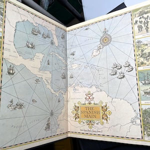 Puede incluir: Libro abierto con un mapa antiguo del Caribe, titulado "The Spanish Main". El mapa presenta ilustraciones de barcos, escenas hist&oacute;ricas y una rosa de los vientos. La paleta de colores incluye azules, beiges y amarillos.
