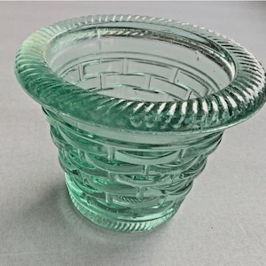 May include: A clear, light green glass basket-weave design container. The container has a wide, textured rim and a basket-like pattern on the sides. The glass has a slight tint, giving it a vintage appearance.