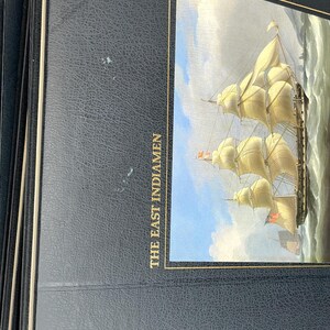 Puede incluir: Una pila de libros de tapa dura azul oscuro. La portada del libro superior presenta una pintura de un velero alto con velas blancas ondeantes sobre un mar agitado. El t&iacute;tulo "THE EAST INDIAMEN" est&aacute; impreso en oro.