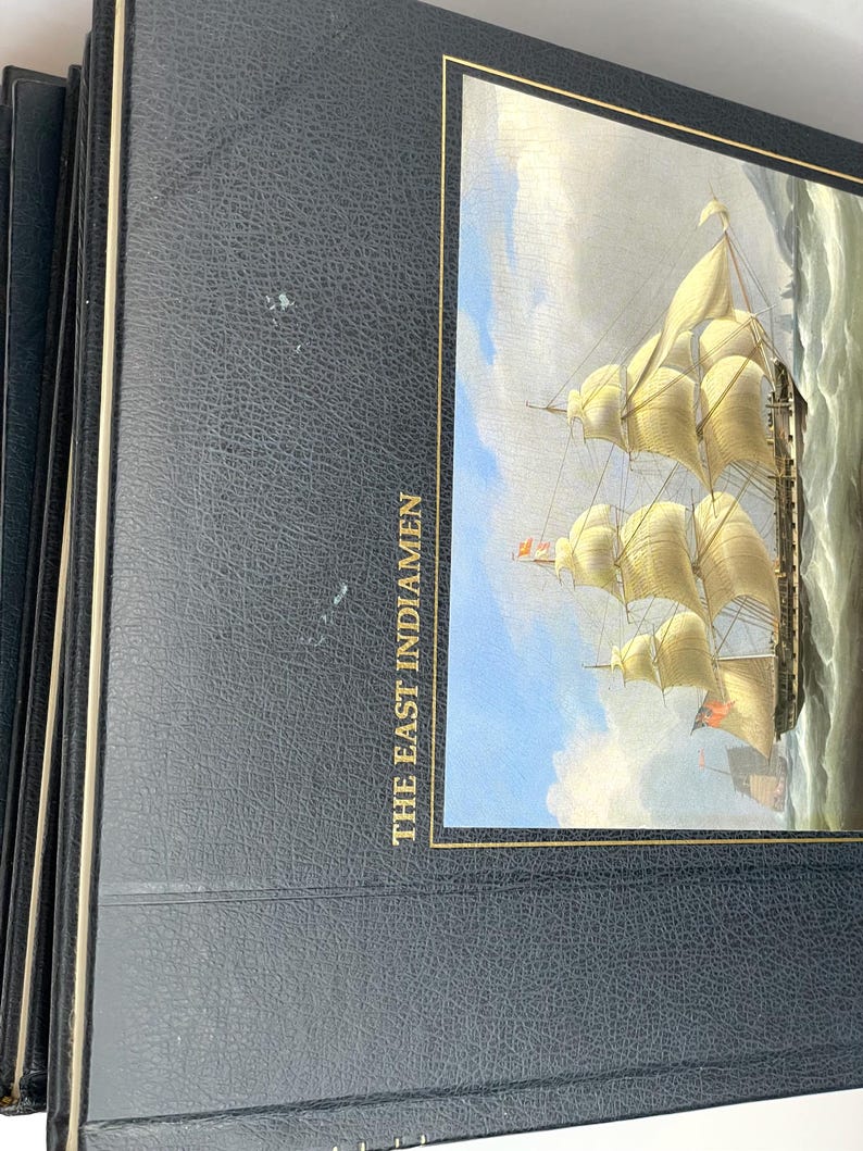 Puede incluir: Una pila de libros de tapa dura azul oscuro. La portada del libro superior presenta una pintura de un velero alto con velas blancas ondeantes sobre un mar agitado. El t&iacute;tulo "THE EAST INDIAMEN" est&aacute; impreso en oro.