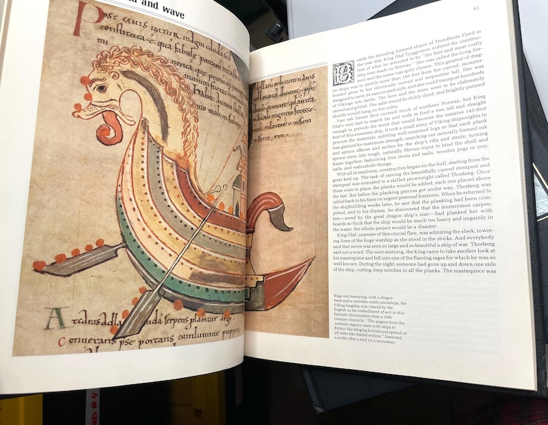 Puede incluir: P&aacute;gina de un libro abierto con una colorida ilustraci&oacute;n de un barco vikingo. El barco presenta una cabeza de drag&oacute;n, remos y un casco a rayas en rojo, amarillo, verde y azul. Texto en lat&iacute;n rodea la imagen.