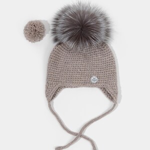 Puede incluir: Un gorro de punto color topo con orejeras y un gran pompón gris y esponjoso en la parte superior. Un pompón más pequeño a juego se encuentra a un lado. El gorro tiene cintas y un pequeño emblema plateado.