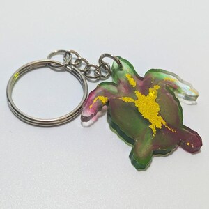 May include: A sea turtle keychain with a silver key ring and chain. The turtle is made of translucent resin with green, pink, and gold accents. The keychain is a decorative accessory.