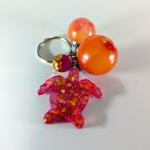 May include: A pink resin turtle keyring with gold glitter embedded in the resin. The keyring has two orange resin balls attached to a silver metal ring.