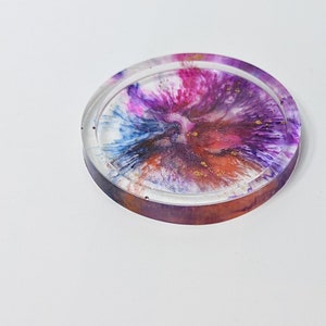 Puede incluir: Un posavasos redondo de resina transparente con un diseño abstracto colorido en tonos azules, rosas, morados y naranjas con destellos dorados.