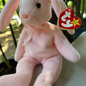 May include: Pink plush bunny rabbit with a white ribbon around its neck. The bunny is sitting on a surface and has a Ty Beanie Baby tag attached to its right ear. The tag says "Beanie Babies Baby".