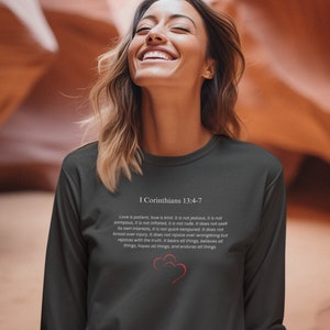 May include: A dark gray long-sleeve shirt with a white print of a Bible verse from 1 Corinthians 13:4-7. The verse is about the characteristics of love. The shirt features a small red heart graphic.