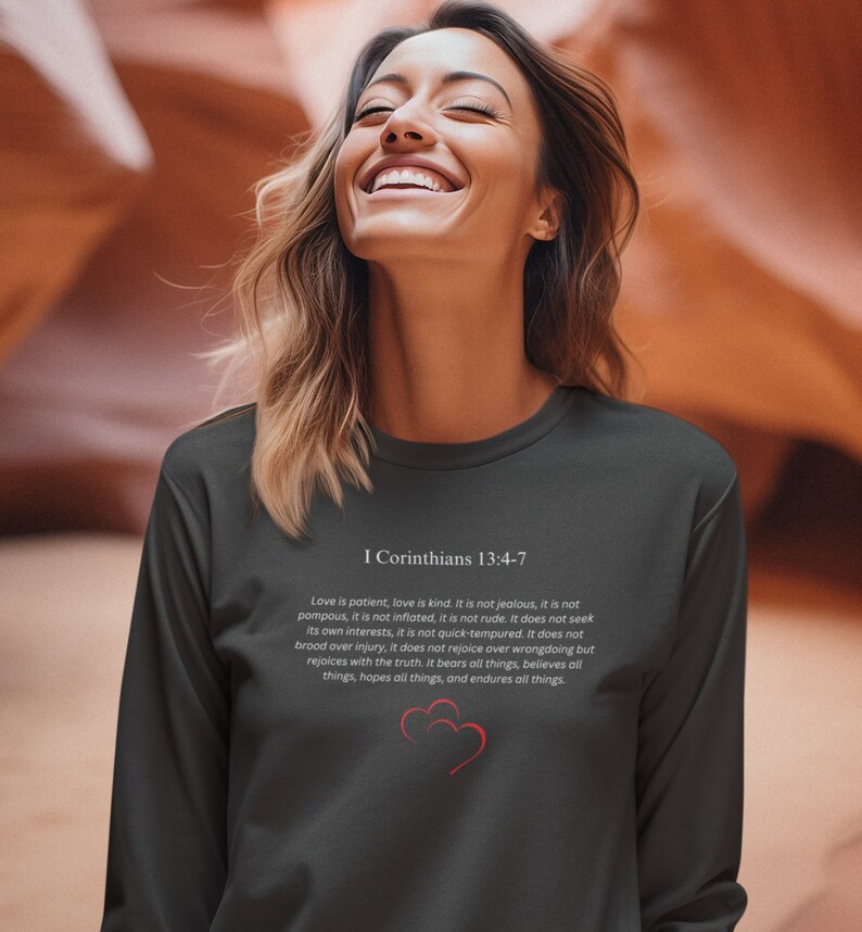 May include: A dark gray long-sleeve shirt with a white print of a Bible verse from 1 Corinthians 13:4-7. The verse is about the characteristics of love. The shirt features a small red heart graphic.
