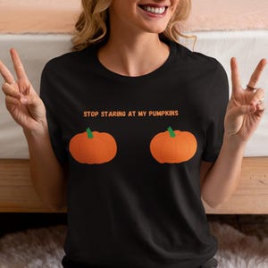 May include: Black t-shirt with the text "STOP STARING AT MY PUMPKINS" in orange. The shirt features two orange pumpkin graphics. The shirt is a fun and playful design, perfect for Halloween or autumn.