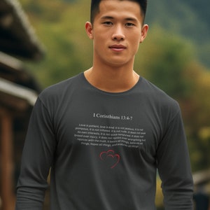 May include: A dark gray long-sleeve shirt with a white print of the Bible verse 1 Corinthians 13:4-7. The verse is about the characteristics of love. The shirt features a red heart graphic.
