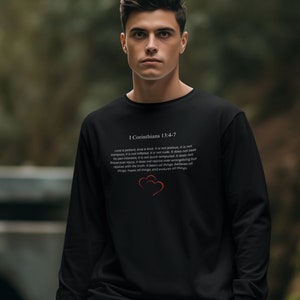 May include: Black long-sleeve sweatshirt with a white print of a Bible verse from 1 Corinthians 13:4-7. The verse is about love being patient, kind, and not jealous. The print also includes two red hearts.