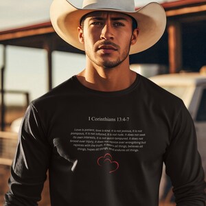 May include: Black long-sleeve shirt with a white print of the Bible verse I Corinthians 13:4-7. The verse is about the characteristics of love. The shirt features a red heart graphic.
