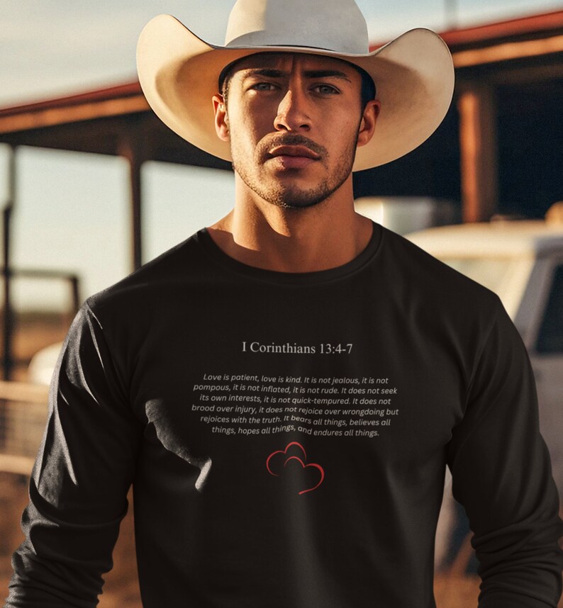 May include: Black long-sleeve shirt with a white print of the Bible verse I Corinthians 13:4-7. The verse is about the characteristics of love. The shirt features a red heart graphic.