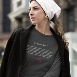 May include: A dark gray long-sleeve t-shirt with a white print of a Bible verse from I Corinthians 13:4-7. The verse is about love being patient, kind, and not jealous. Two red hearts are printed below the verse.