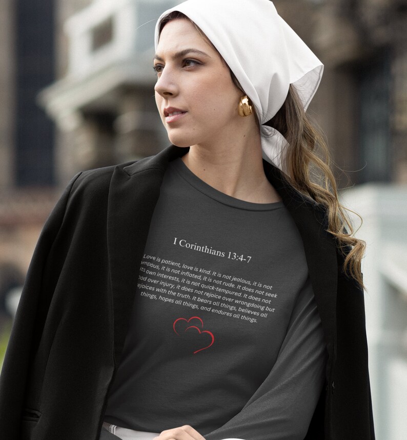 May include: A dark gray long-sleeve t-shirt with a white print of a Bible verse from I Corinthians 13:4-7. The verse is about love being patient, kind, and not jealous. Two red hearts are printed below the verse.