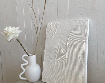 May include: White textured canvas wall art with a raised circular pattern. The art is leaning against a white wall next to a white vase with a white flower.